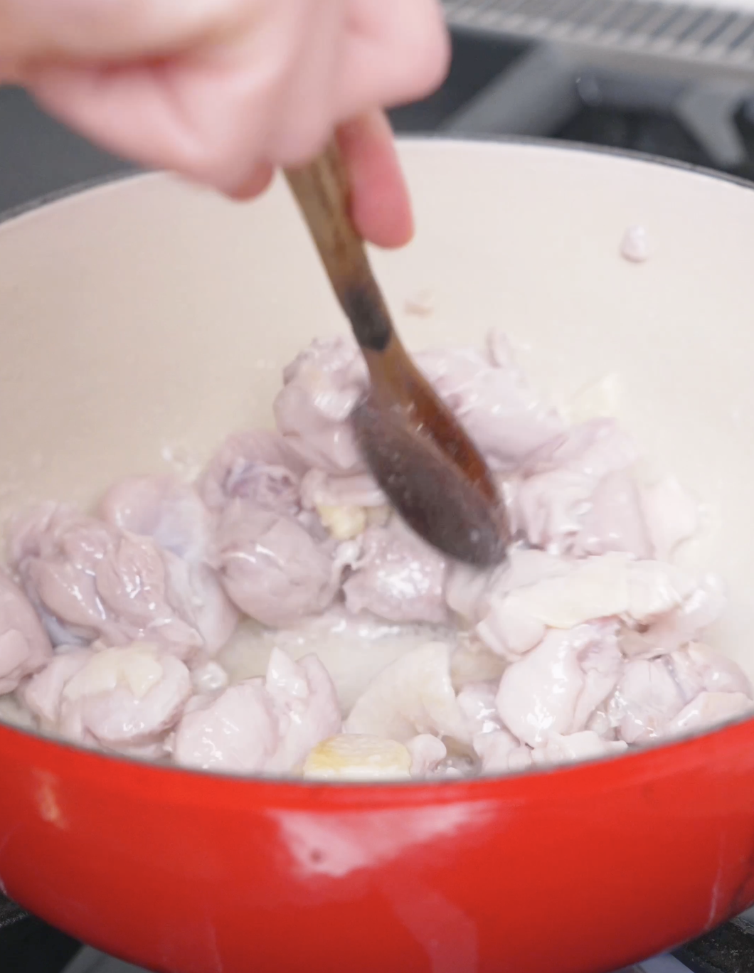 Cubed chicken thighs being stirred with a wooden spoon in a red dutch oven for Chicken Curry Stew Recipe.