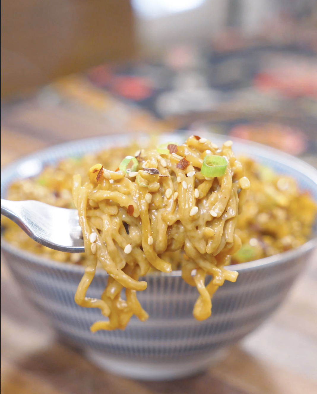 A fork lifting a twirl of sauce-coated noodles with sesame seeds and chili flakes above the bowl of Creamy Curry Noodles Recipe.