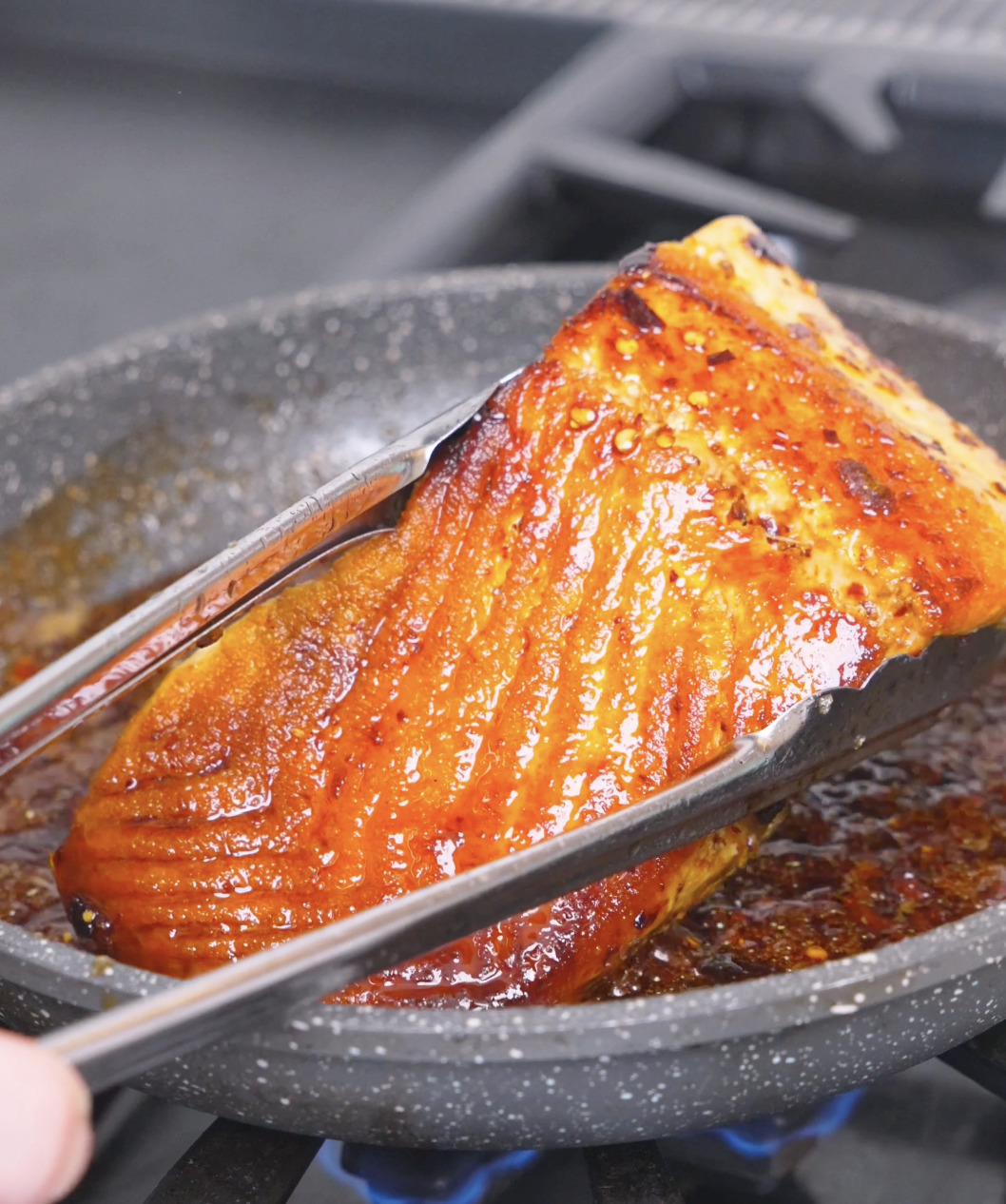 Tongs lifting the salmon as the sauce thickens into a deep red glaze for Spicy Salmon Bowl Recipe.