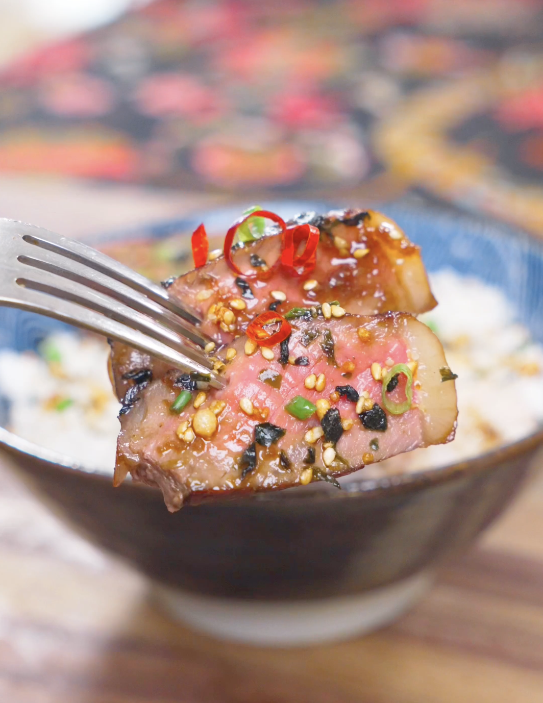 A fork lifting two medium-rare slices topped with furikake and red pepper above the bowl of Japanese Steak Bowl Recipe.