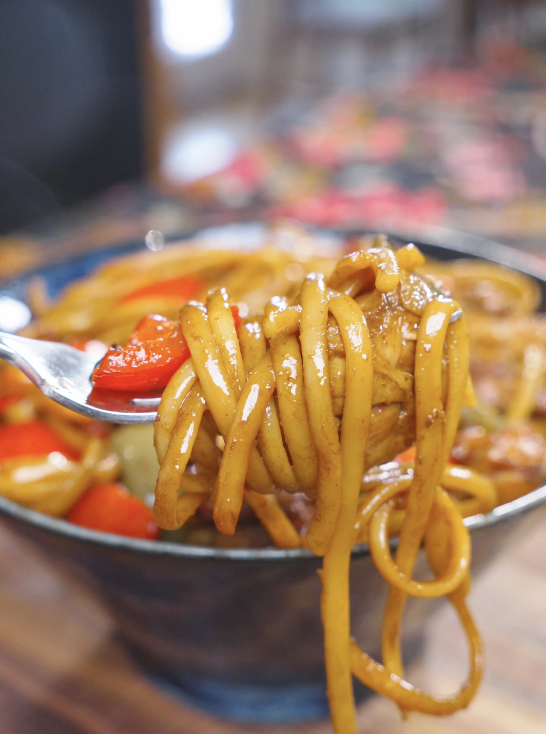 A fork lifting a twirl of glossy noodles with chicken and red pepper above the bowl of Kung Pao Chicken Noodles Recipe.