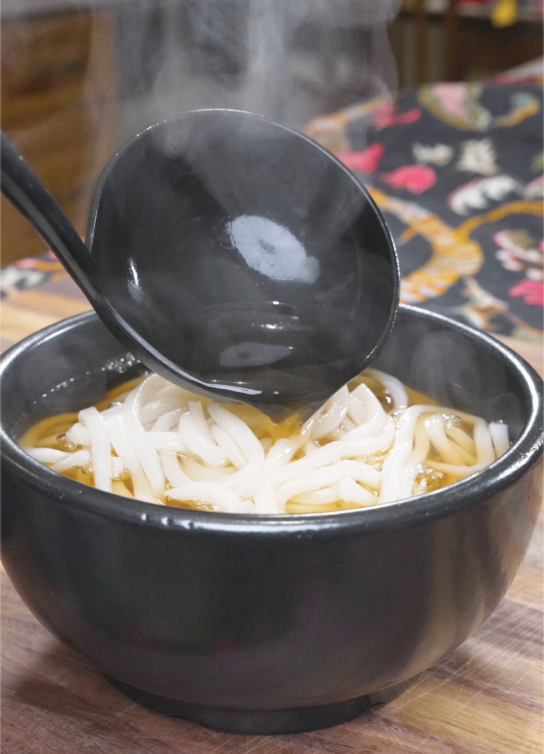 Steaming dashi broth being ladled over udon noodles in a black bowl for Beef Udon Recipe.