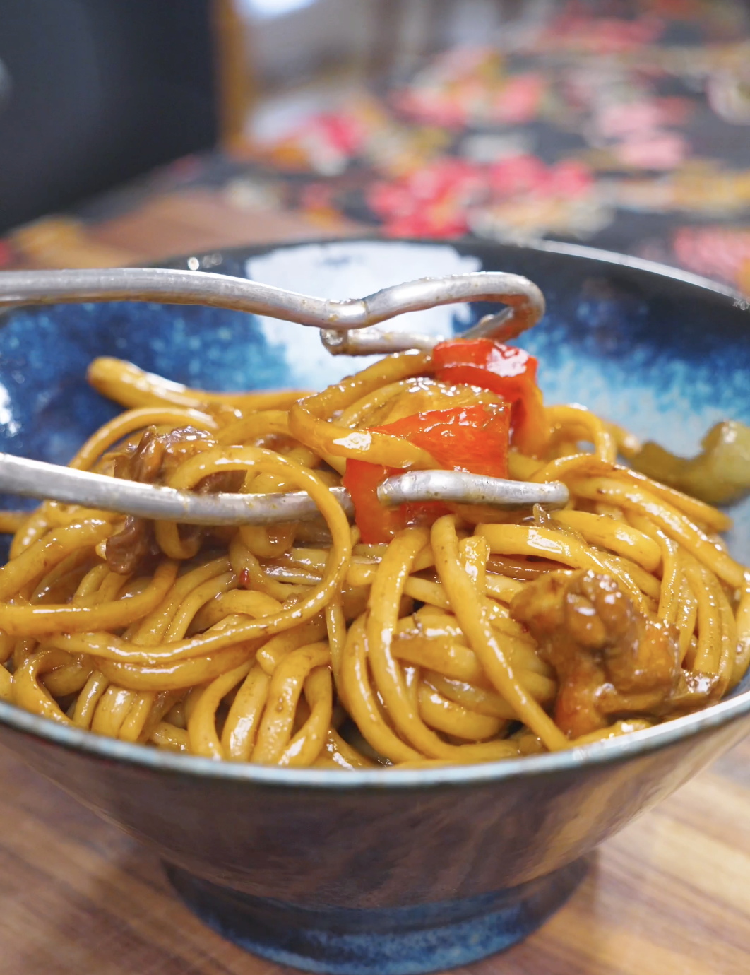 Tongs placing a tangle of sauced noodles and peppers into a blue bowl for Kung Pao Chicken Noodles Recipe.