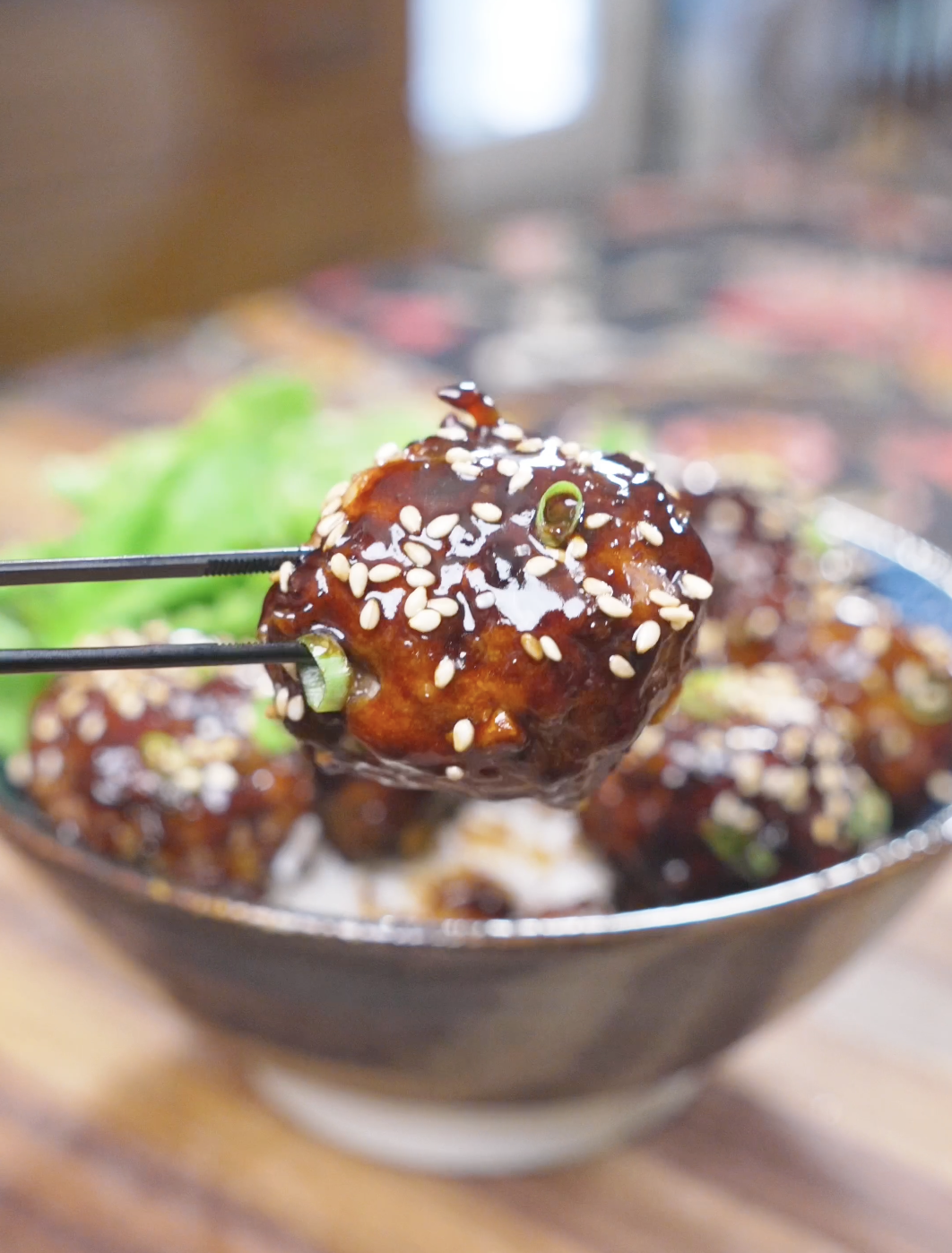 Chopsticks holding a single lacquered, sesame-covered meatball above the bowl of Teriyaki Meatballs Recipe.