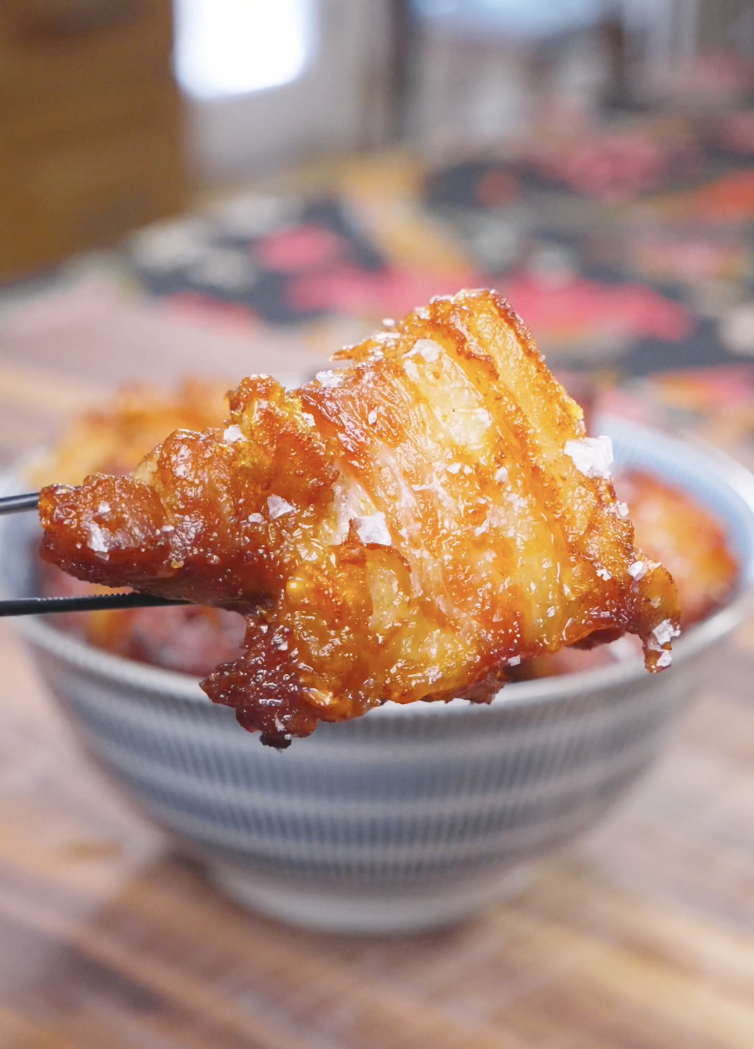 Chopsticks holding a crisp-crusted cube with a soft center above the bowl of Deep Fried Pork Belly Recipe.