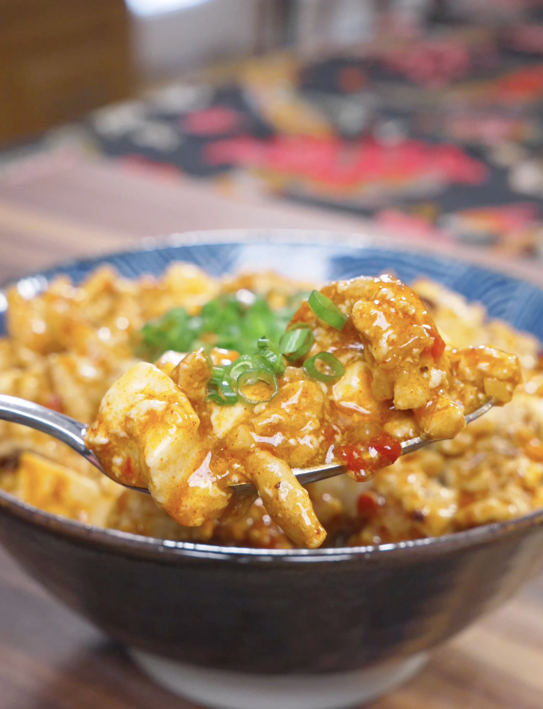 A spoon lifting soft tofu and pork in glossy red sauce above the bowl of Mapo Tofu Recipe.