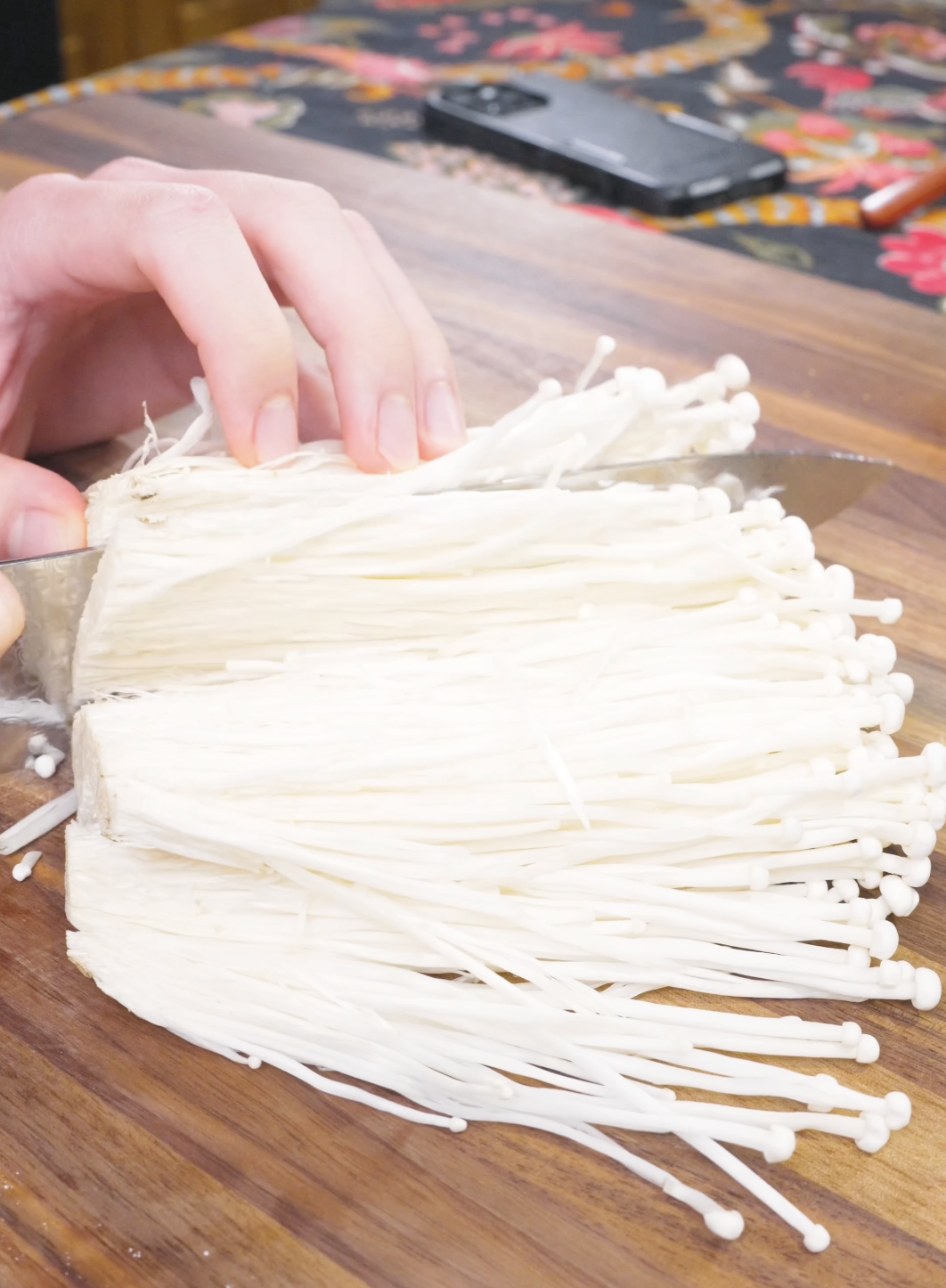 A cluster of enoki mushrooms being cut thinly lengthwise on a wooden board for Crispy Fried Enoki Mushrooms Recipe.